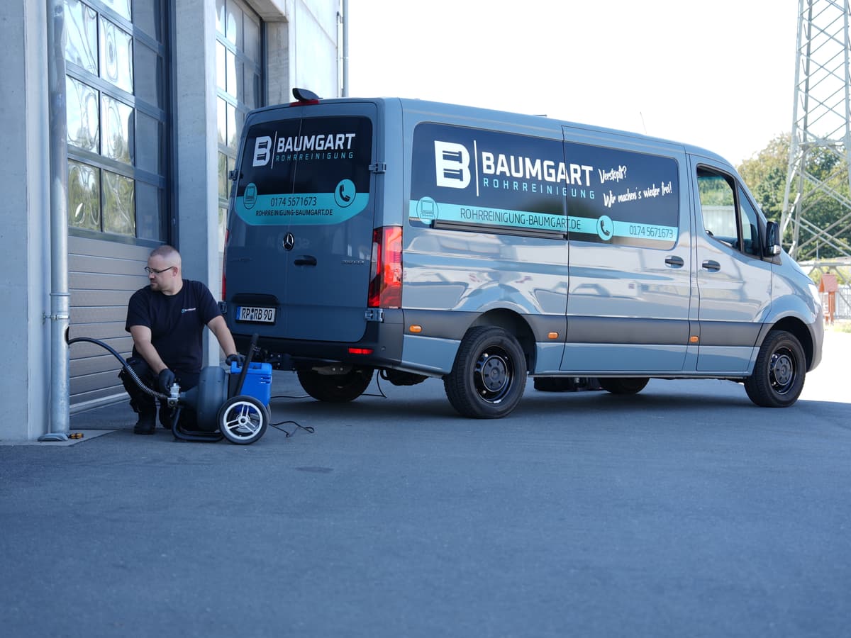 Monteur mit Motorspirale an einem Fallrohr, dahinter das Servicefahrzeug von Rohrreinigung Baumgart